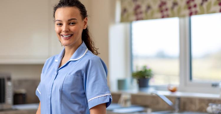 Waist up portrait of a nurse dressed in her scrubs uniform standing indoors at home, looking at the camera with a cheerful expression on her face.