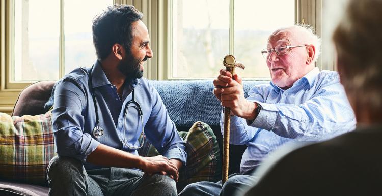 Young male doctor and elderly man sitting on sofa and smiling during care home visit