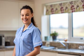 Waist up portrait of a nurse dressed in her scrubs uniform standing indoors at home, looking at the camera with a cheerful expression on her face.